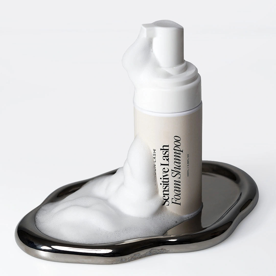 London Lash Sensitive Lash Foam Shampoo displayed on a reflective silver tray with rich white cleansing foam flowing around the bottle.